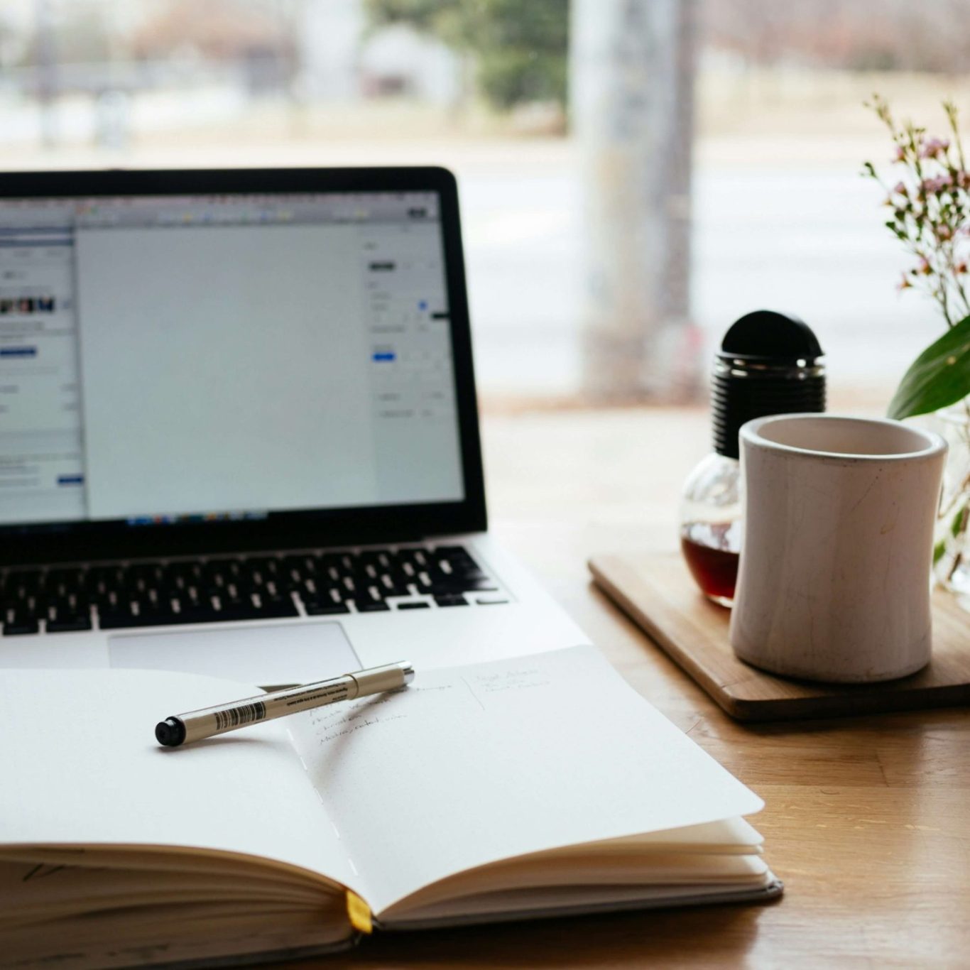 Meet the Expert Behind the Service A laptop, an open notebook, a pen, a coffee mug, and a plant on a wooden table.